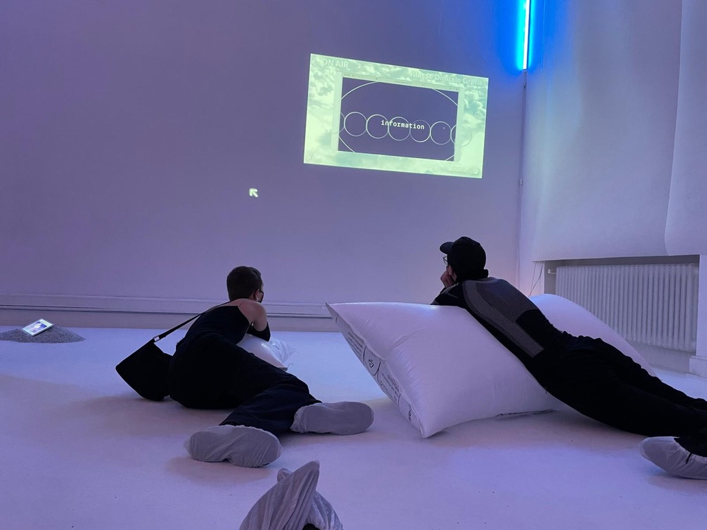 In a white room, big cloudy pillows are tumbling on the ground. One wall shows a video projection.