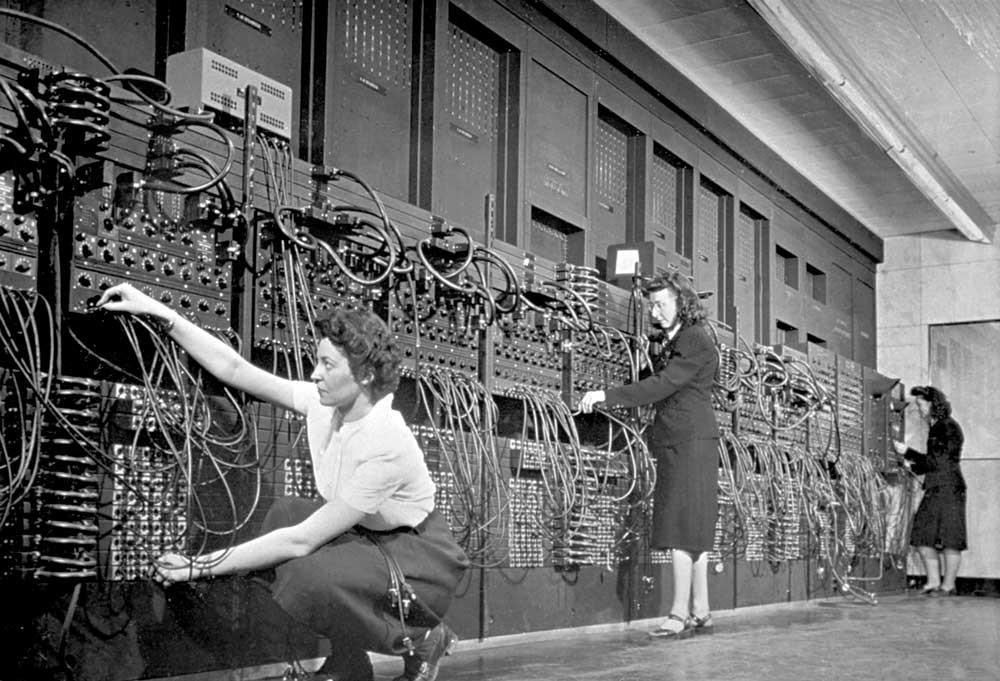 Women standing behind a roomsized machine, plucking cables that stick out of it.