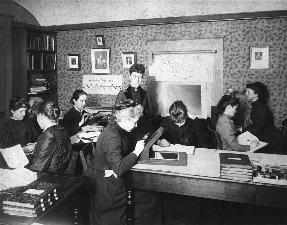 An old Black and White Photograph of eight women sitting in a small room. There Are a lot of Books on Tables and the womens laps.